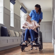 Senior Woman At Home Being Pushed In Wheelchair By Female Care Worker In Uniform